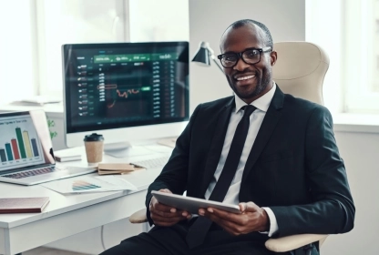 Black trader working in an office, analyzing trading charts on a computer screen.
