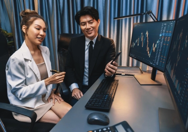 Asian man and woman traders sitting in an office, viewing trading charts on a computer screen.