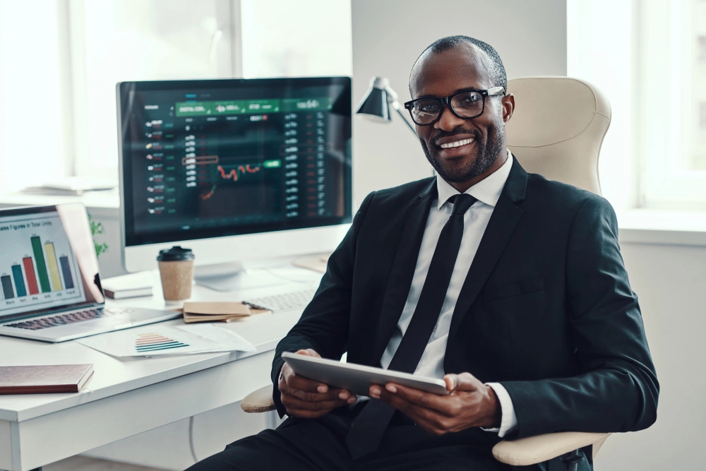 Black trader working in an office, analyzing trading charts on a computer screen.