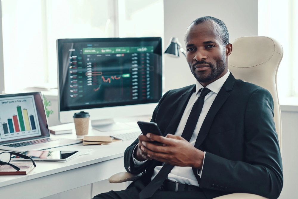 Black trader working in an office, analyzing trading charts on a computer screen.
